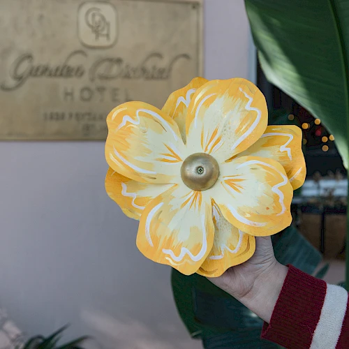 A person holds a bright yellow flower with a circular center in front of a hotel sign and green leaves, sunset lights in the background.