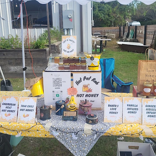 A small outdoor market stall with boxes labeled hot sauces and a centerpiece sign, jars and a samplers table under a canopy.