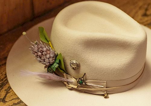 A white fedora with a decorative lavender sprig and small pins, resting on a wooden surface.