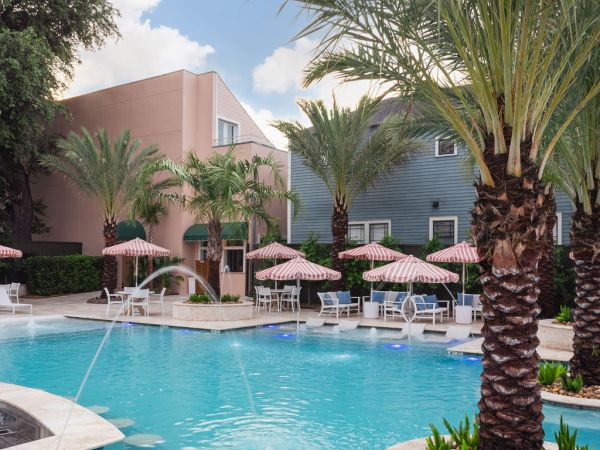 A sunny poolside scene with palm trees, blue water, lounge chairs, beige umbrellas, and pastel buildings in the background, inviting relaxation.
