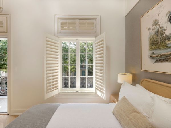 A bright hotel bedroom with a large open window, white shutters, a cozy bed, soft lighting, and a framed landscape painting above the headboard.