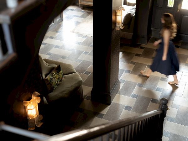 A girl in a blue dress walks through a tiled hall near a stairwell, with a lit lamp and seating area on the left, in a home.