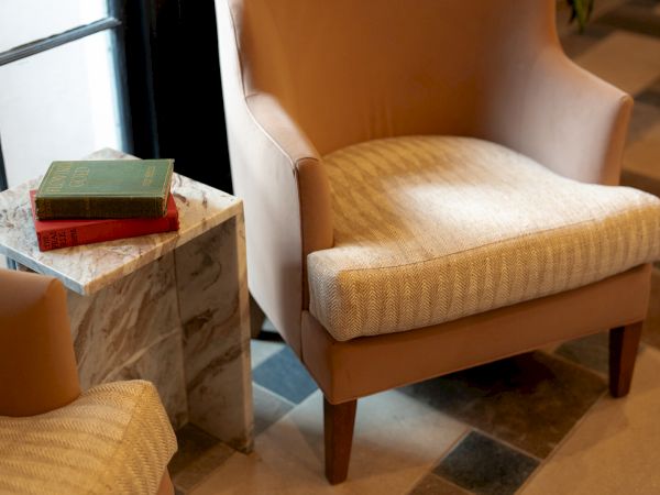 A cozy hotel lobby corner with two padded armchairs, a marble side table stacked with books, and natural light from a window.