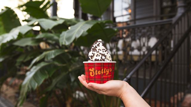 A hand holds a red cup labeled &ldquo;Bettys&rdquo; with a scoop of cookies and cream ice cream topped with chocolate chips, outdoors near greenery.