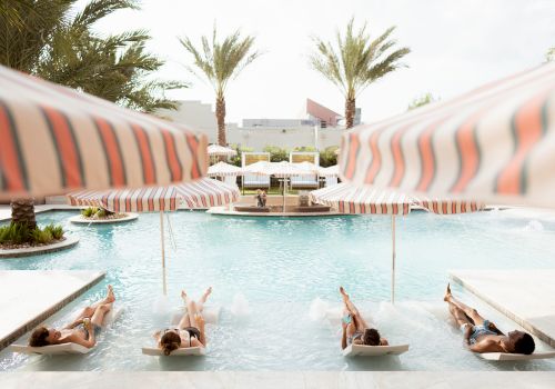 People lounging on pool loungers under striped umbrellas around a sunny pool, palm trees in the background.