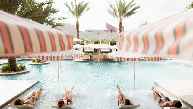 People lounging on pool loungers under striped umbrellas around a sunny pool, palm trees in the background.