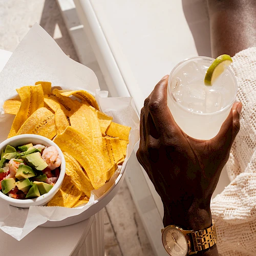 A person lounging with a snack platter: tortilla chips and a bowl of salsa/guacamole, plus a refreshing drink with a lime wedge, relaxation vibes.