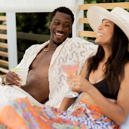 Two friends lounge by the pool, smiling and chatting; a man holds a drink while a woman in a hat sips from a glass, enjoying sunny vibes.