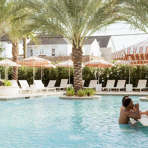 A sunny pool scene with palm trees, lounge chairs, striped umbrellas, and people swimming and chatting by the water.