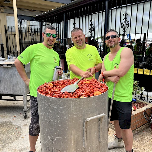 Three men in neon shirts stir a giant pot of saucy meat, cooking outdoors for a crowd, with smiles and teamwork.