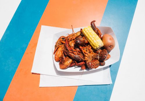 A plate of fried seafood with corn on the cob and a small bun, on layered white napkins placed on a colorful striped background.
