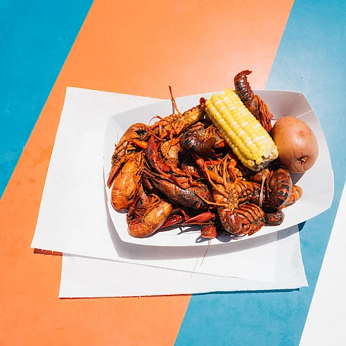A plate of fried seafood with corn on the cob and a small bun, on layered white napkins placed on a colorful striped background.