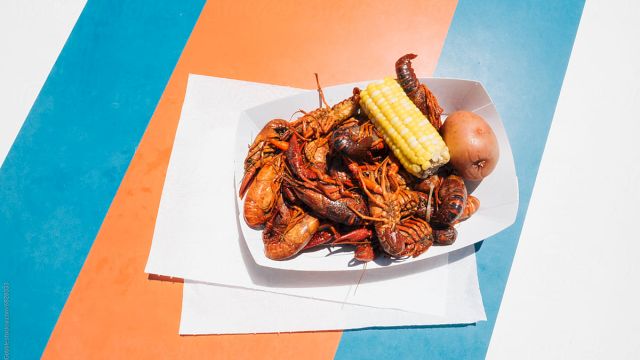 A plate of fried seafood with corn on the cob and a small bun, on layered white napkins placed on a colorful striped background.