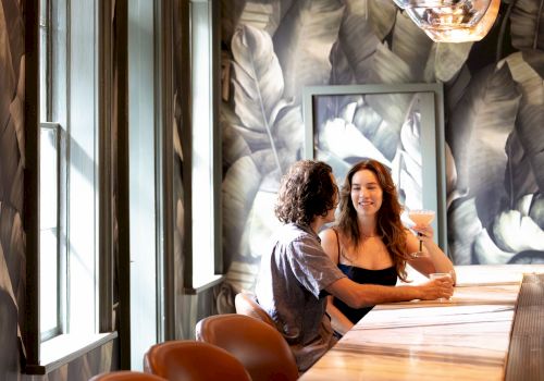 Two people sit at a long wooden bar by a window, chatting and smiling in a stylish, leaf-patterned restaurant interior.