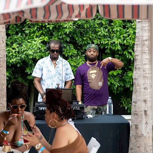 Two DJs spin records under a striped canopy while a small crowd chat and sip nearby, outdoors with greenery behind them.
