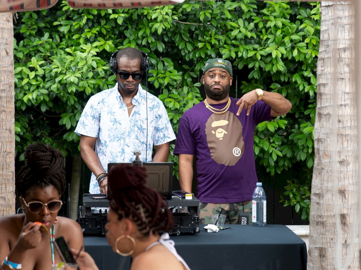 Two DJs playing at an outdoor party under a striped canopy, with guests seated nearby and lush greenery in the background, enjoying the vibe.