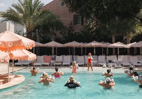 A sunny outdoor pool event where swimmers wearing white water-splash devices play with a crowd of people under striped umbrellas.