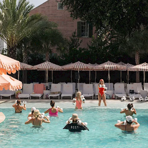 A sunny outdoor pool event where swimmers wearing white water-splash devices play with a crowd of people under striped umbrellas.