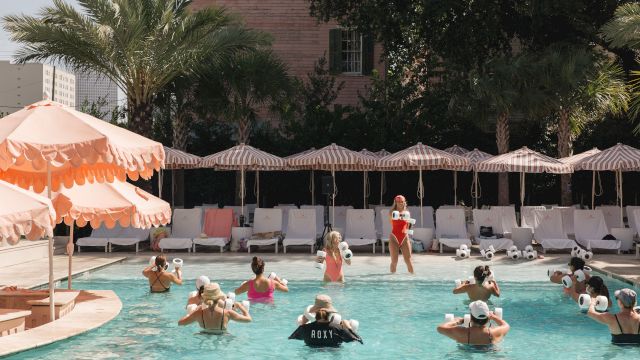 A sunny outdoor pool event where swimmers wearing white water-splash devices play with a crowd of people under striped umbrellas.