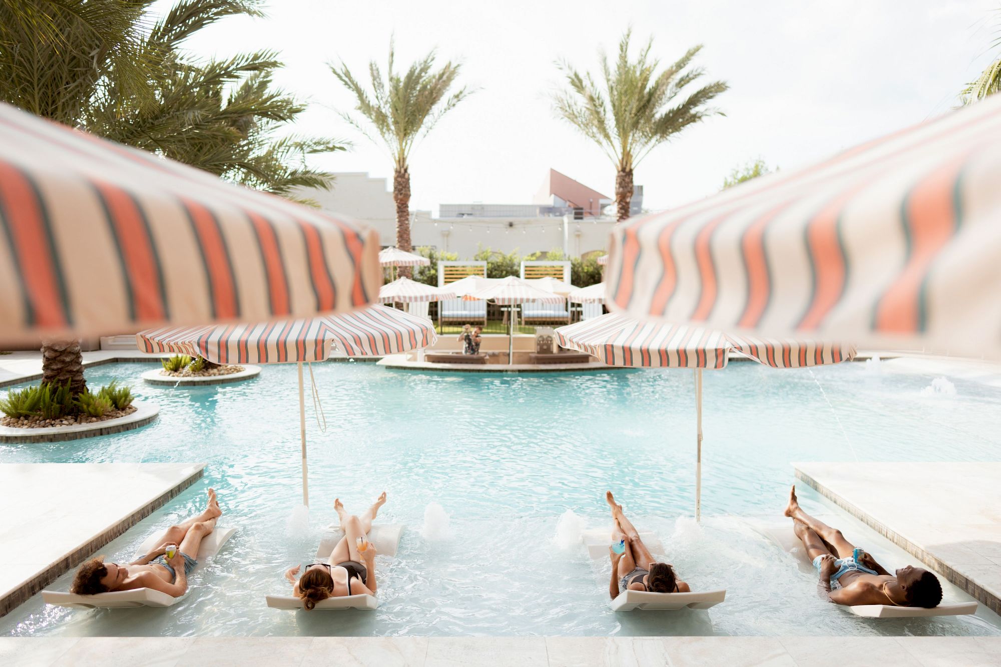 People lounge on submerged sunbeds in a pool under striped umbrellas, relaxing as palm trees frame a resort setting.