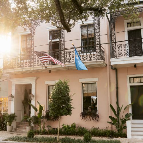 A stately brick townhouse with white trim, arched windows, columned entry, and surrounding trees along a sunny street.