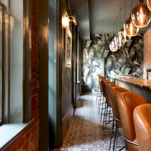 A narrow, stylish bar with a patterned tile floor, dark walls, hanging globe lights, and brown leather stools along a long counter.