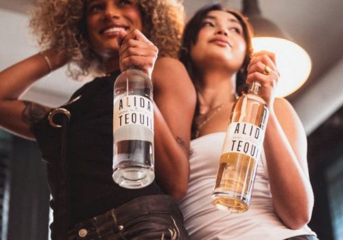 Two women smile while holding tequila bottles labeled &ldquo;ALID TEQUIL&rdquo; at a party.