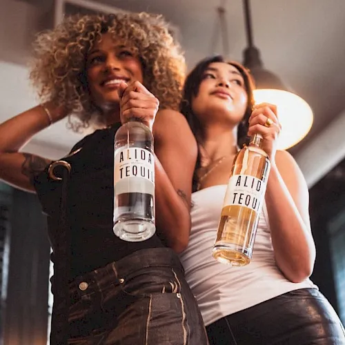 Two women smile while holding tequila bottles labeled &ldquo;ALID TEQUIL&rdquo; at a party.