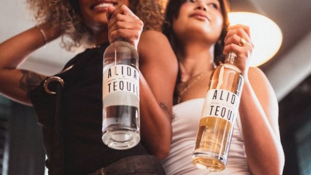 Two women smile while holding tequila bottles labeled &ldquo;ALID TEQUIL&rdquo; at a party.