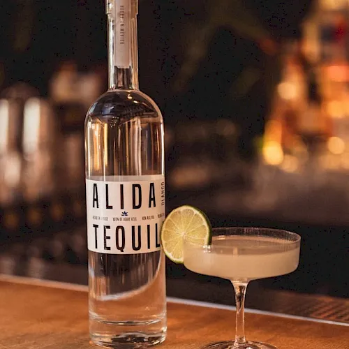 A bottle of Alida tequila sits on a bar beside a lime garnish in a cocktail glass, with blurred bottles in the background.