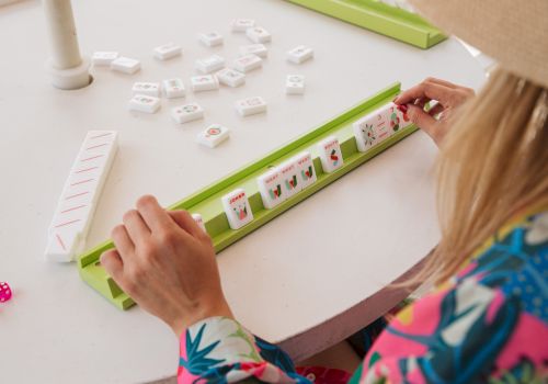 A person&rsquo;s hands are placing small game tiles/cards along a green board in a tabletop board game, with other tiles scattered around.