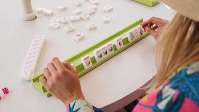 A person&rsquo;s hands are placing small game tiles/cards along a green board in a tabletop board game, with other tiles scattered around.