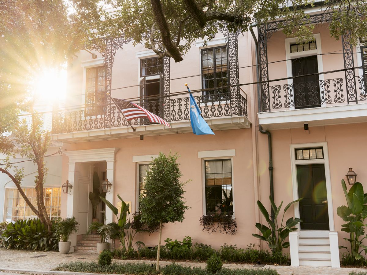 A sunlit courtyard with a white building, wrought-iron balcony railings, two hanging flags, and lush plants and trees in view.