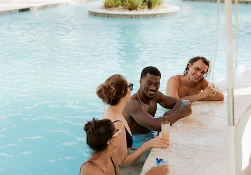 A group of friends relax and laugh by a resort pool, holding drinks and chatting together on a sunny day.