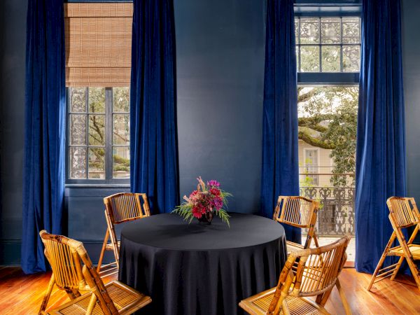 A round table with a black cloth and flower centerpiece sits between bamboo chairs, framed by blue curtains and a window view.