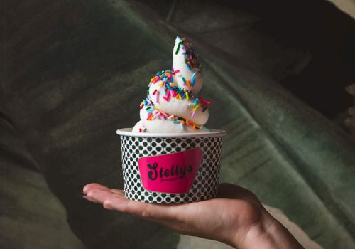 Hand holds a small cup of frosted dough balls/ice-cream topped with colorful sprinkles from J. Elly&rsquo;s, on a couch.