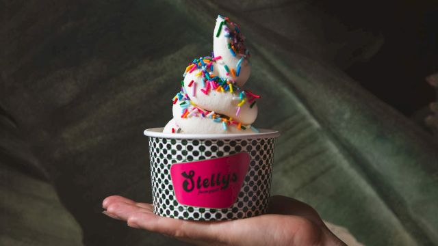 Hand holds a small cup of frosted dough balls/ice-cream topped with colorful sprinkles from J. Elly&rsquo;s, on a couch.
