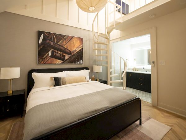 A modern bedroom with a black bed, two nightstands, framed wall art, and a spiral staircase beside an open bathroom vanity.