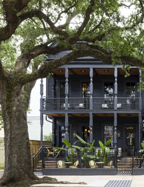 A large leafy tree towers over a gray two-story porch with ornate railings and house number 1612, creating a shady entrance.