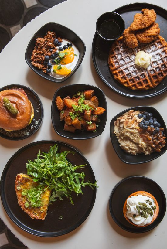 A spread of breakfast/brunch dishes: waffles with butter, fried chicken, yogurt, granola oats, and a sandwich on plates.