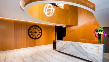 A modern lobby with a marble desk, circular wooden design overhead, large clock on the wall, and a vase of flowers.