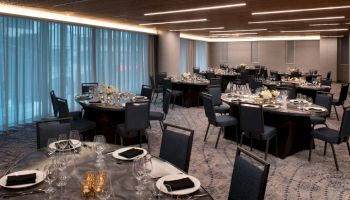 The image shows an elegantly set banquet room with round tables, chairs, plates, glasses, and floral centerpieces under a modern ceiling.