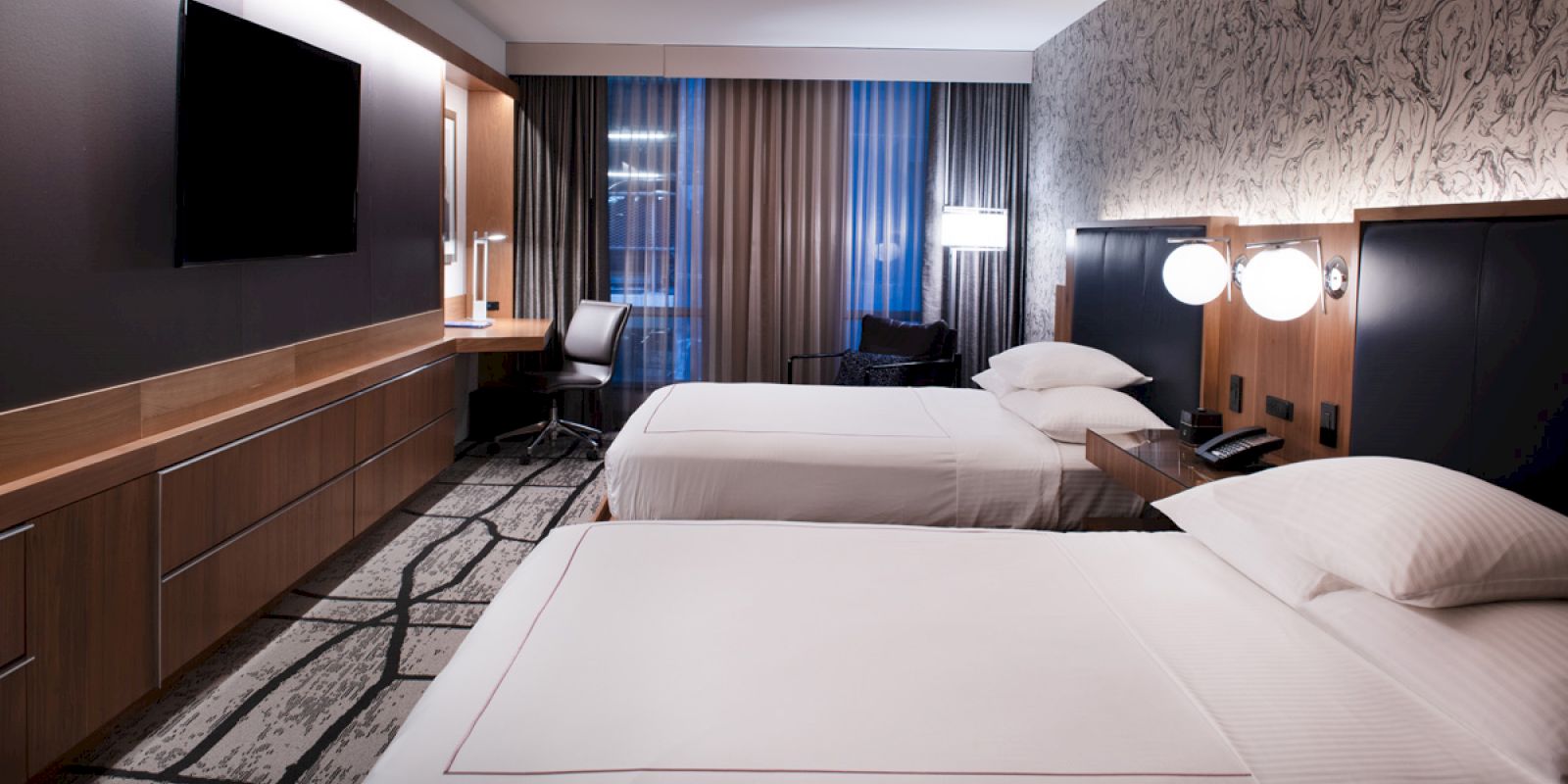 The image shows a modern hotel room with two beds, a large TV, a desk with a chair, and a stylish carpet and decor.