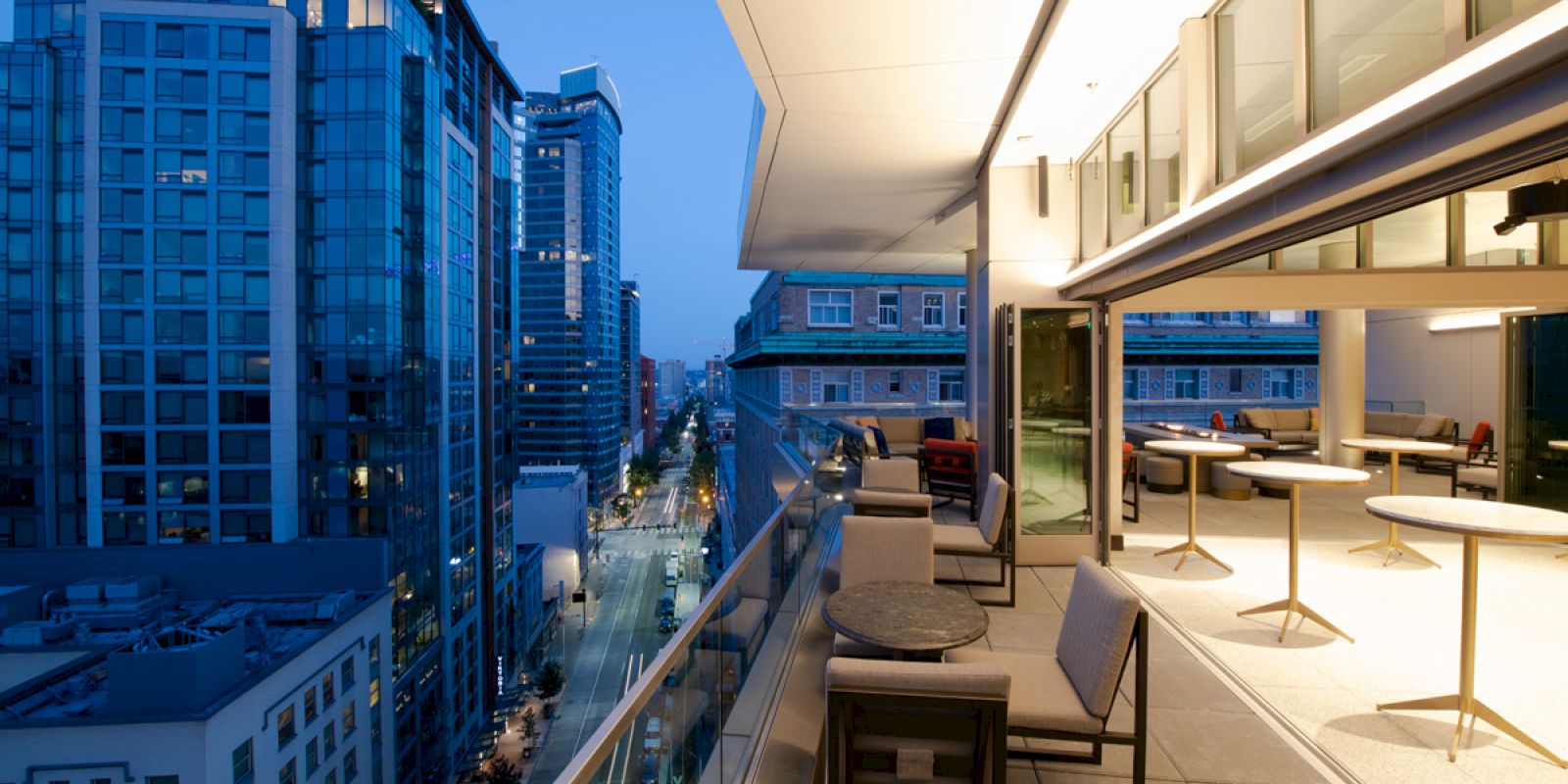 A modern urban balcony overlooking a city street at dusk, featuring outdoor seating and glass railings.