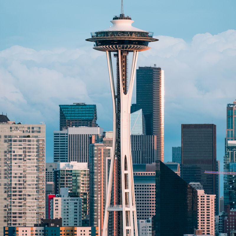 The image shows the Space Needle in Seattle, surrounded by tall buildings against a backdrop of a cloudy sky and cityscape.