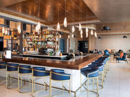 This image shows a modern bar with blue chairs, hanging lights, shelves of bottles, and a cozy seating area in the background.