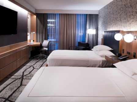 The image shows a modern hotel room with two neatly made beds, a TV, a desk, and a window with curtains allowing natural light.