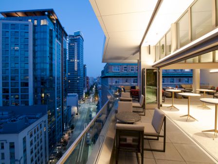 A modern balcony with seating overlooks a city street lined with tall buildings during evening.