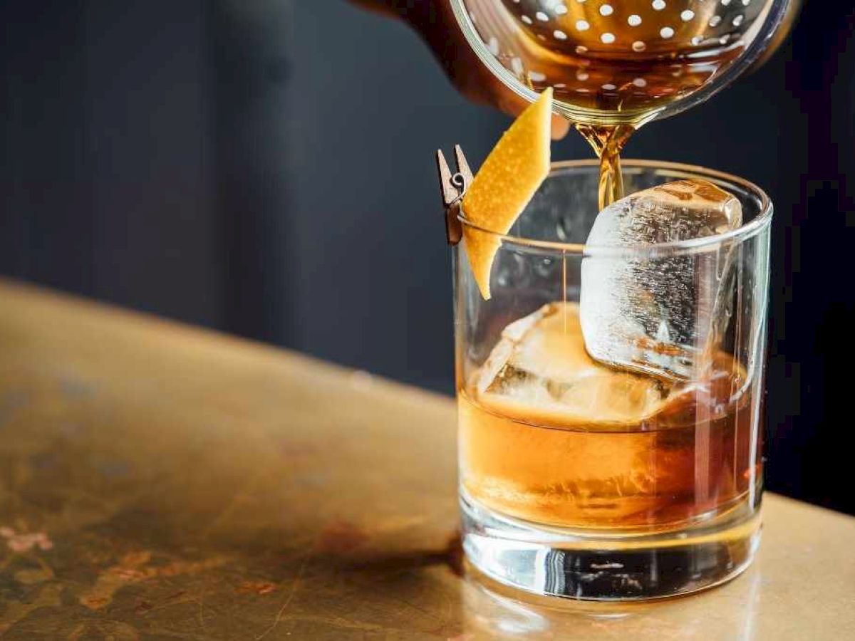 A cocktail is being poured into a glass with ice and a lemon twist garnish on a bar.