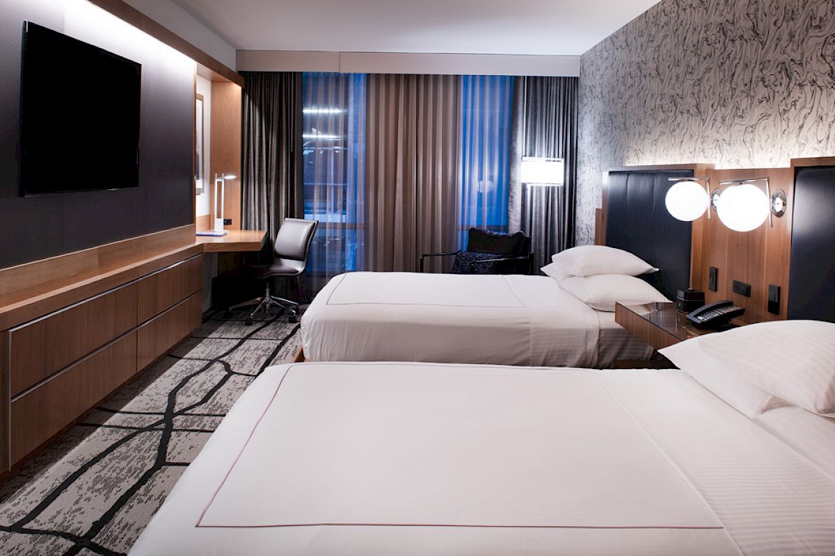 The image shows a modern hotel room with two neatly made beds, a TV, desk, chair, and stylish lighting.